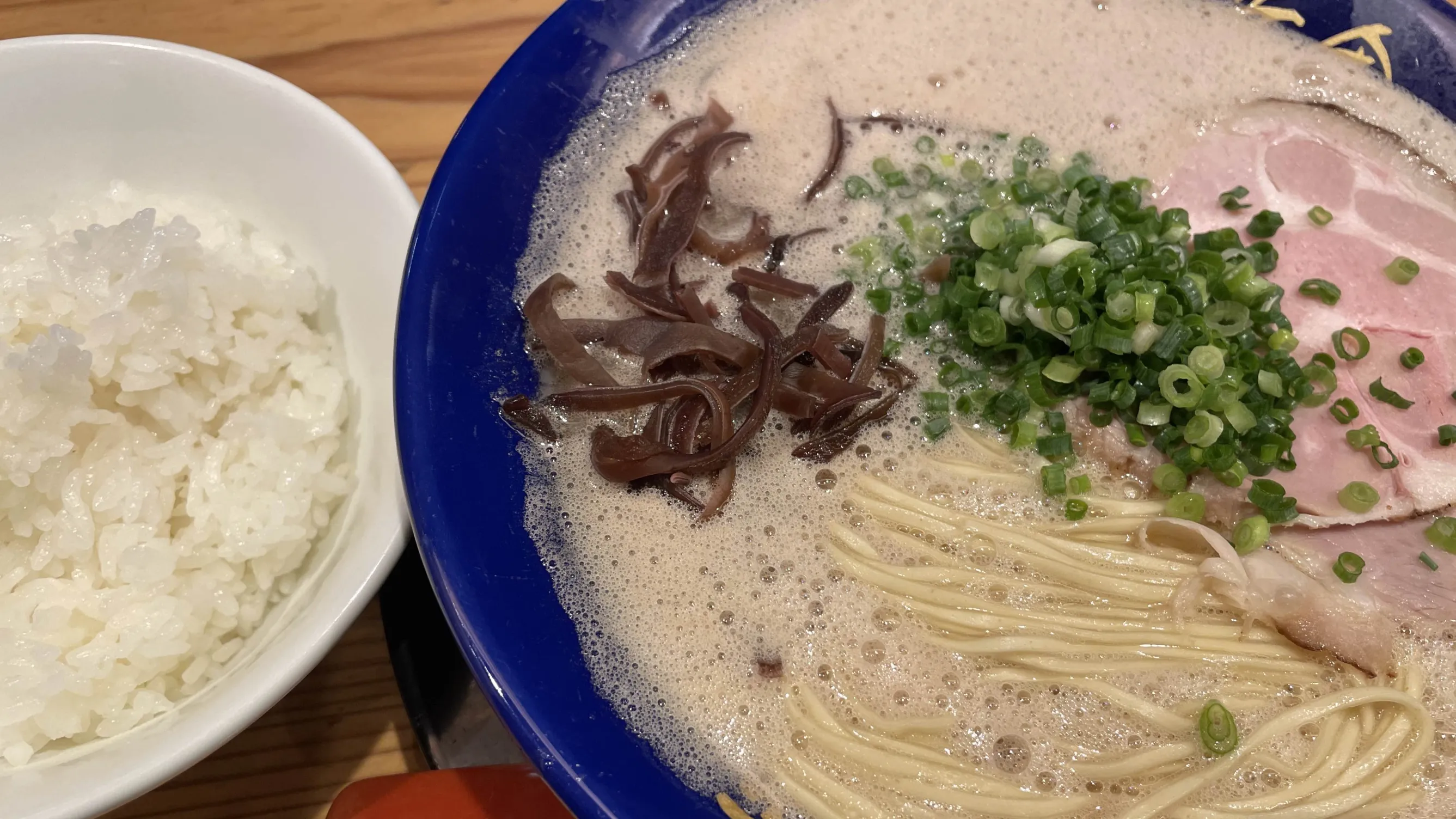 「博多一幸舎 総本店」の「泡系豚骨ラーメン」のバリカタとライスの写真。きくらげ、刻みネギ、チャーシューがトッピングされ、スープには泡が浮かんでいる。